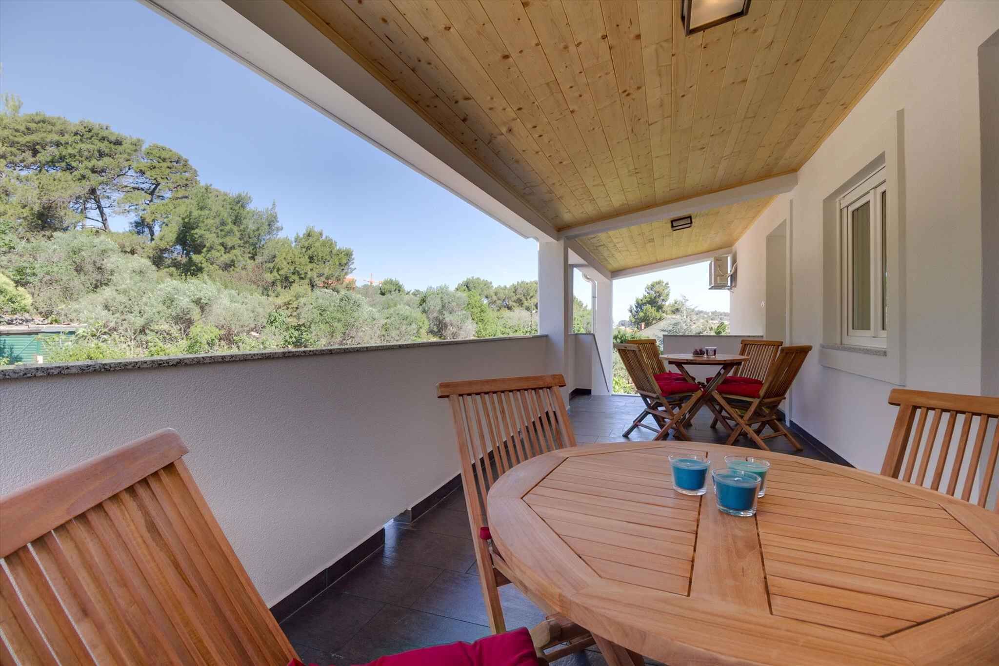 Ein überdachter Außenbereich mit Holztisch und Stühlen, Blick auf Bäume und einen Pool im Hintergrund.