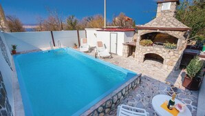 Una piscina con suelo de piedra, una cocina al aire libre de construcción de piedra y una silla blanca.