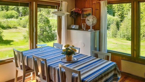 Zona pranzo con tavolo ricoperto da tovaglia a righe, circondato da sedie di legno. Sul tavolo, un vaso con fiori gialli e, sopra uno scaffale, una ventola.
