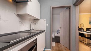 Eine moderne Küche mit schwarzem Arbeitsplatz, weißen Schränken und einer Marmor-Spritzschutzwand. Der Boden ist aus Holz und eine Tür führt in einen Essbereich mit Tisch und Stühlen.
