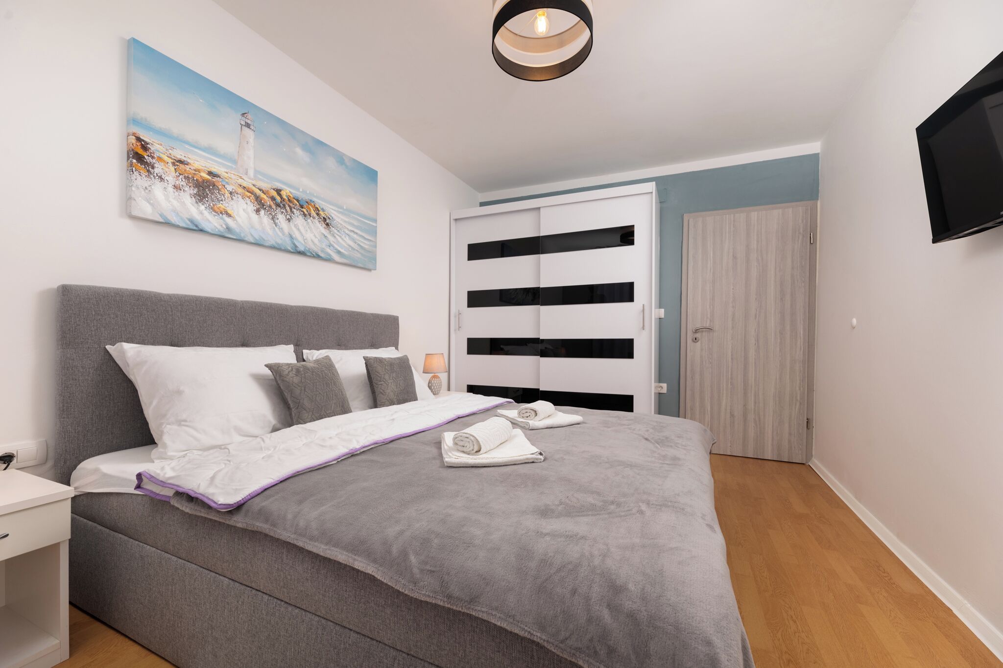 A modern bedroom with a grey bed, white pillows, a painting of a lighthouse, a TV mounted on the wall, and a wooden floor.