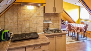 Une cuisine compacte équipée d’une cuisinière, d’un évier et de meubles en bois. On aperçoit à l’arrière-plan un coin repas avec une table et des chaises à carreaux.