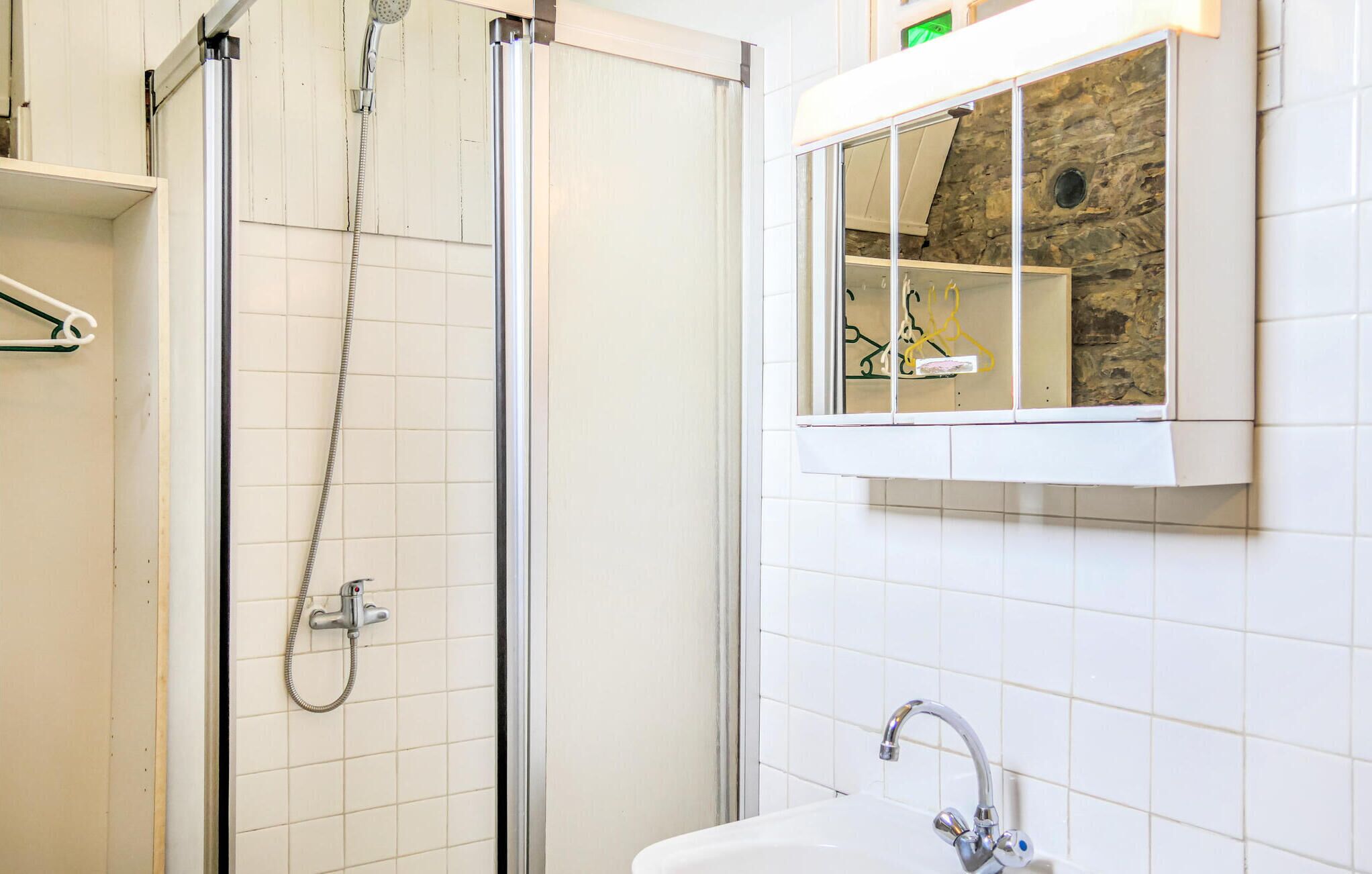 Ducha con puerta de cristal, pared de azulejos blancos y ventana con un muro de piedra al fondo.