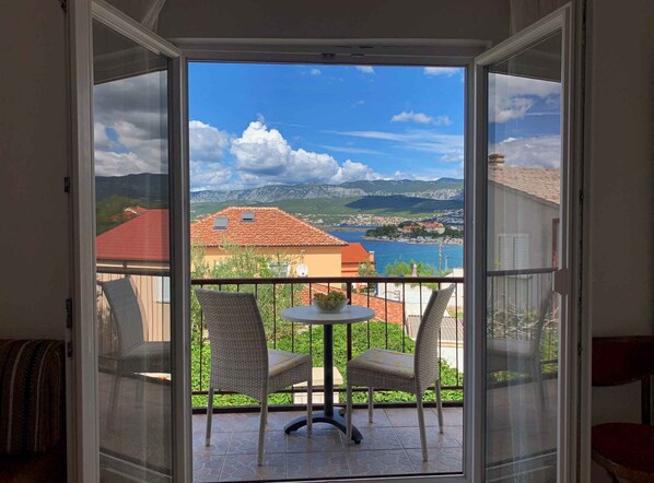 A balcony with two wicker chairs and a small table, overlooking a scenic view of a lake, mountains, and a town.