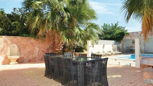 An outdoor patio with a glass table surrounded by wicker chairs, a stone wall, and a pool in the background. - Nice home in Le Grau d'Agde (Le Grau d'Agde)