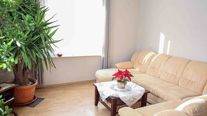 Un salon avec un canapé beige, une table basse en bois recouverte d’une nappe à motifs floraux, une plante en pot et une fenêtre avec des rideaux.