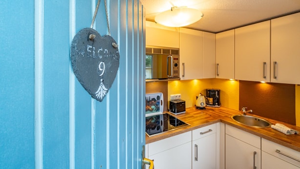 A kitchen with white cabinets, a wooden countertop, a sink, and a hanging heart-shaped decoration with the word "WELCOME". - App.9 - Priel Hüs, Germany (List)