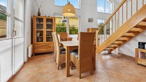 A spacious dining area with a wooden table and wicker chairs, a wooden cabinet with glass doors, and a staircase with wooden railings. - Beach front home in Süssau/Ostsee (Süssau/Ostsee)