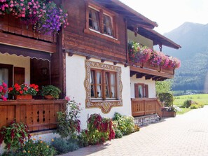 Una casa de madera tradicional con balcones adornados con flores, un camino de piedra y montañas al fondo.