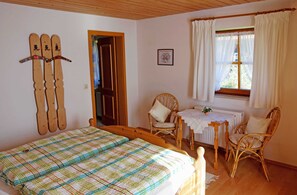 Une chambre à coucher avec un lit, deux fauteuils en osier, une petite table, un radiateur et une fenêtre avec des rideaux.
