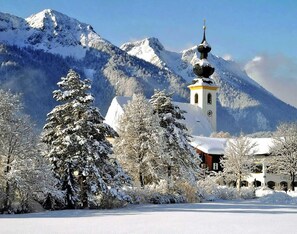 Una iglesia cubierta de nieve con un campanario, rodeada de árboles cargados de nieve y montañas al fondo.