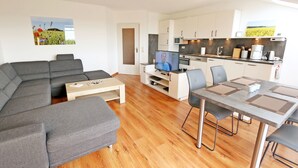 A modern living room with a grey sectional sofa, a wooden coffee table, and a flat-screen TV. A dining area with a table and chairs is adjacent to the kitchen, which has white cabinets and a microwave. - F: SEEMANN Apartment Whg. C14 with balcony - SEEMANN Apartment Whg. C14 with balcony - SEEMANN Apartment Whg. (Thiessow)