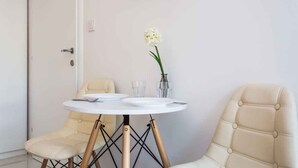 Un pequeño comedor con una mesa redonda blanca, dos sillas color beige, un florero con una flor blanca y un vaso.