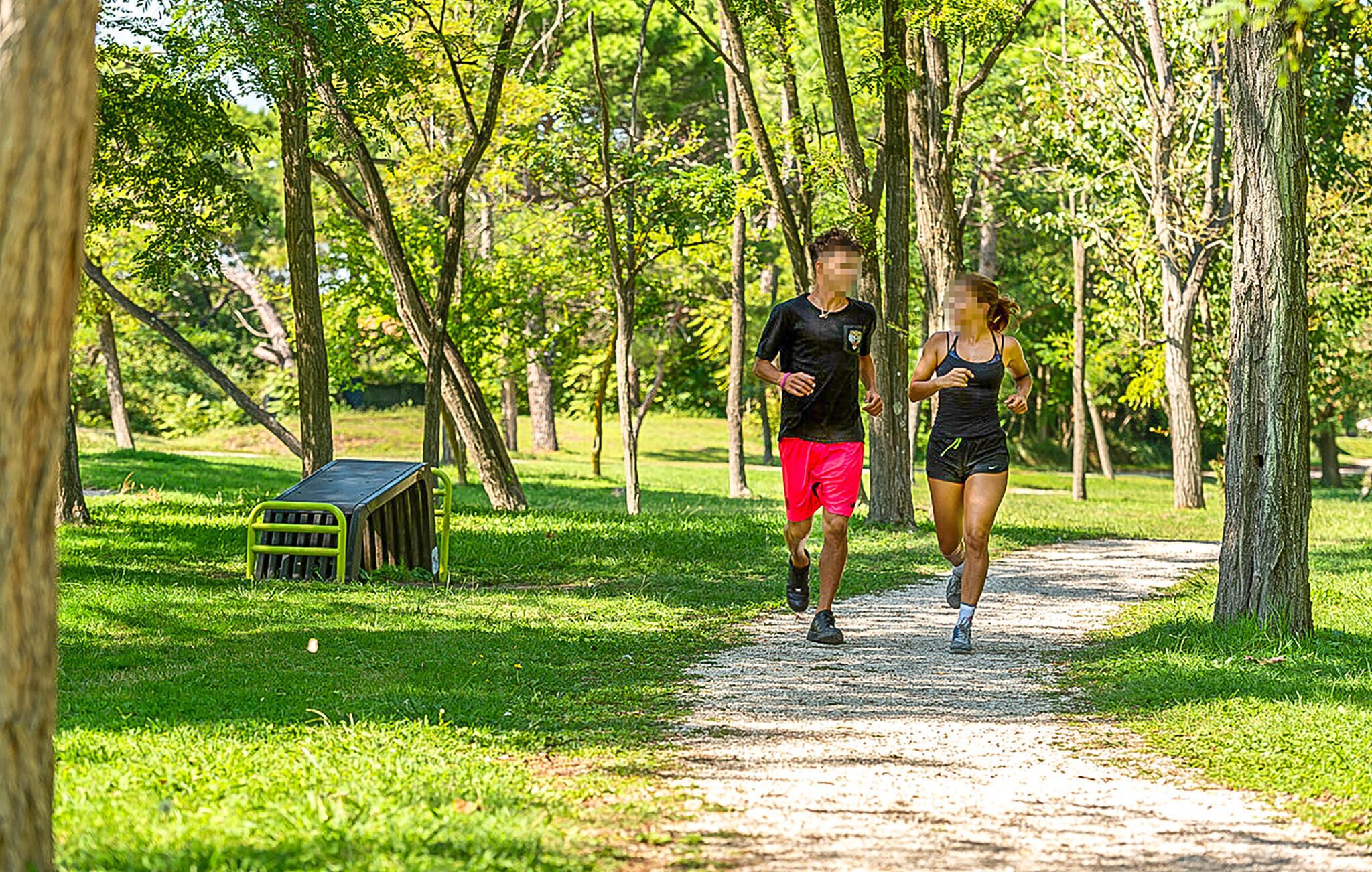 Zwei Personen joggen auf einem Kiesweg in einem Park mit Bäumen und einer kleinen Gartenhütte.