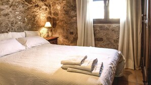A bedroom with a stone wall, a bed with white bedding, a nightstand with a lamp, and a window with curtains. - Stunning home in Villanueva del Duque (Villanueva del Duque)