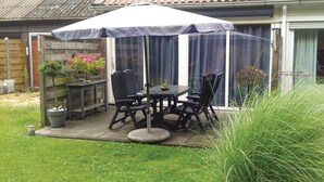 Eine Terrasse mit Tisch und Stühlen unter einem Sonnenschirm, umgeben von Kübelpflanzen und einem Holzzaun.