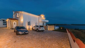 A modern two-story house with a stone driveway, two cars parked in front, and a view of the sea at dusk.