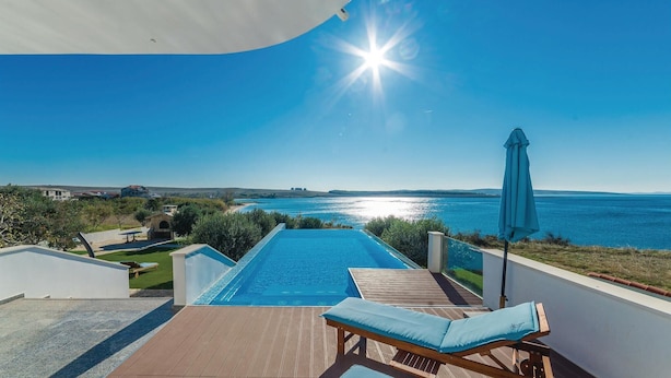 A poolside view with a clear blue sky, a sunlit pool, and a view of the sea.