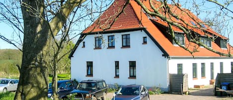 En hvid bygning med rødt tag, parkerede biler og et træ i forgrunden.