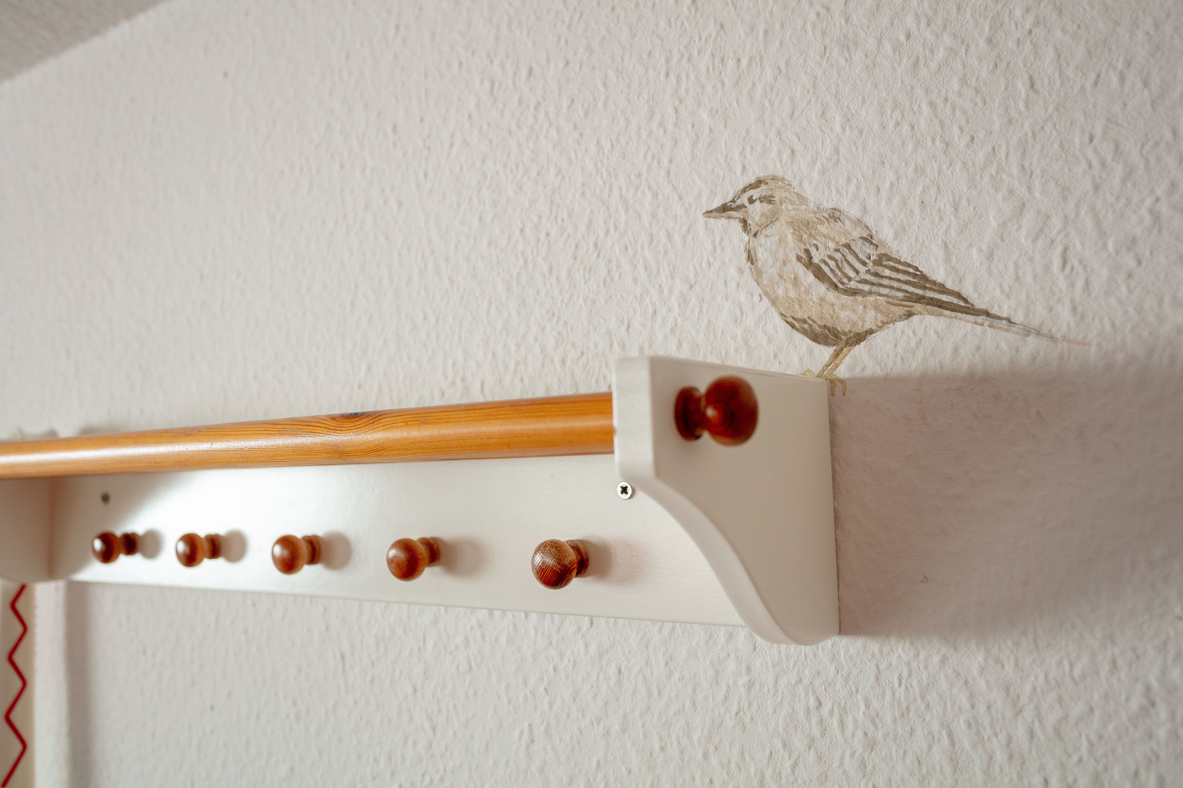 Eine an der Wand befestigte Holzkonstruktion mit einer Vogelillustration, einer Holzstange und mehreren Rundgriffen.