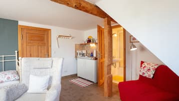 A room with a bed, a red sofa, a white armchair, a wooden ceiling beam, a kitchen area, and a bathroom.