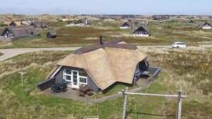 A thatched-roof house with a patio area, surrounded by a rural landscape with other similar houses. - Gorgeous home in Ringkøbing with sauna (Ringkøbing)