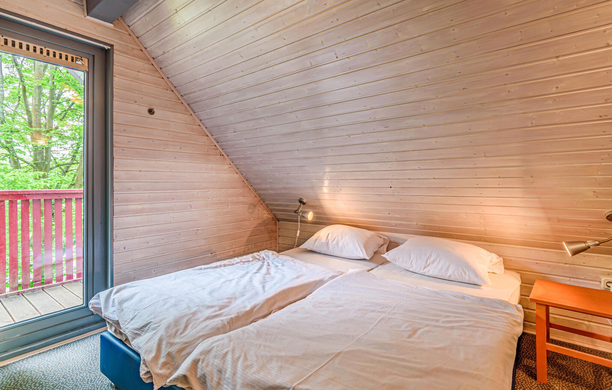 Une chambre avec un plafond en pente, un lit recouvert de draps blancs, une table de chevet et une fenêtre donnant sur des arbres.