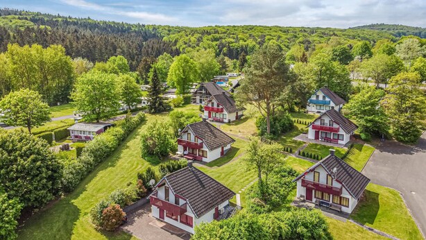 A residential area with multiple houses, each featuring a distinct architectural style with red and white exteriors, surrounded by lush greenery and a hilly backdrop.