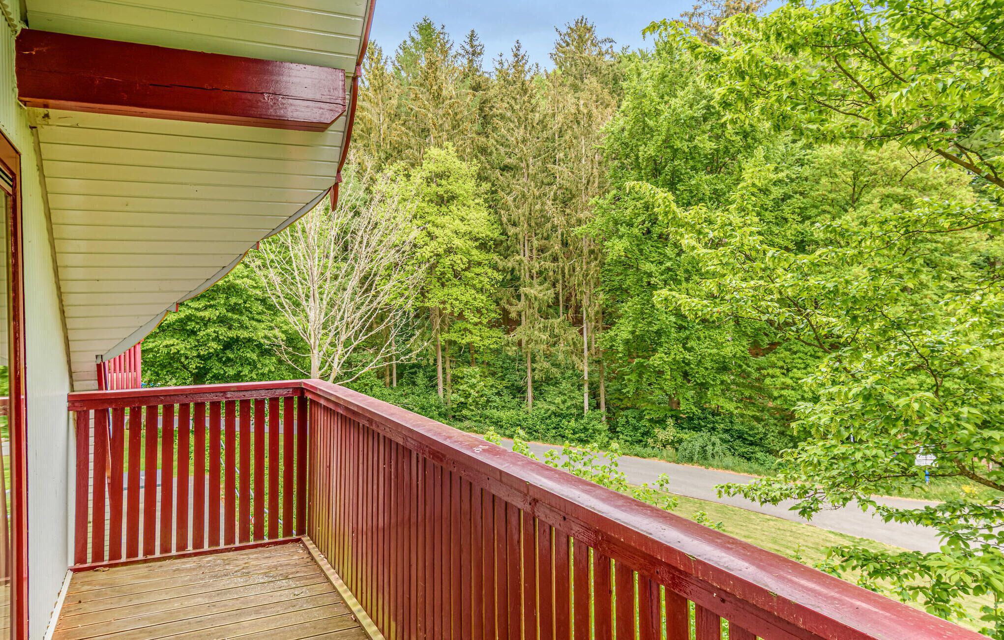 A wooden deck with a red railing overlooking a road and dense green forest.