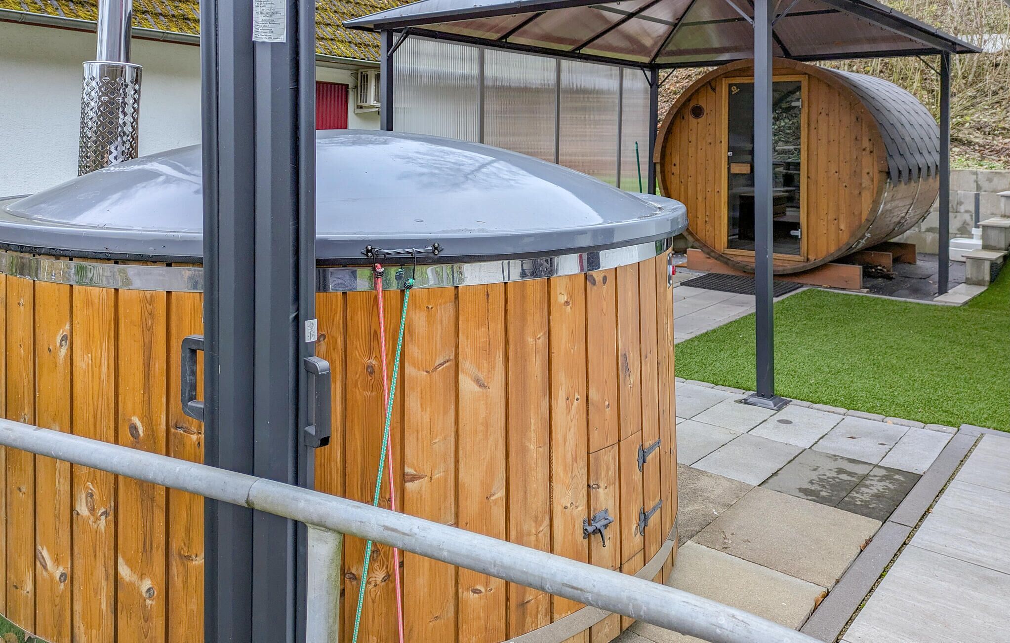 A wooden sauna structure with a metal roof, a metal pole, and a wooden barrel in the background.