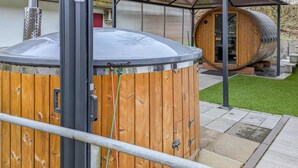 A wooden sauna structure with a metal roof, a metal pole, and a wooden barrel in the background.