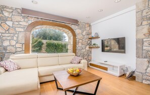 A living room with a stone wall, a white sofa, a wooden coffee table with fruit, and a TV on the wall. - Beautiful home in Malinska (Malinska)
