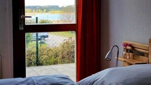 Quarto com cama, janela com vista para a água e vegetação, e um banco de madeira com abajur.