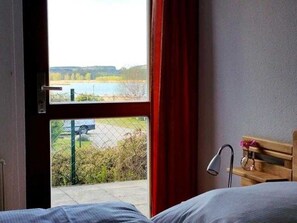 A room with a bed, a window with a view of a body of water and greenery, and a wooden bench with a lamp.