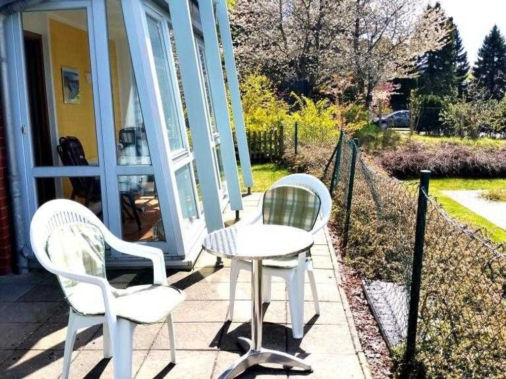 A patio with two white chairs and a small table, a yellow building with a glass door, and a fenced garden area.
