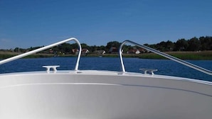 Proa de barco com grades de metal e vista de um lago calmo, com casas ao fundo.