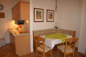 A compact kitchen with a microwave, sink, and wooden cabinets. There is a dining area with a table and chairs, and two framed pictures on the wall.