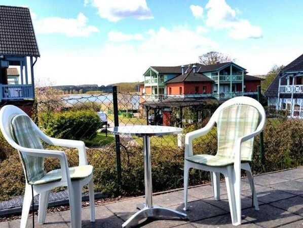 Two white plastic chairs and a small round table on a paved area with a view of houses and a fence in the background.