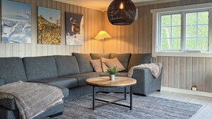 En hyggelig stue med en grå sofa, et rundt træbord og et stort vindue med udsigt til grønne omgivelser.