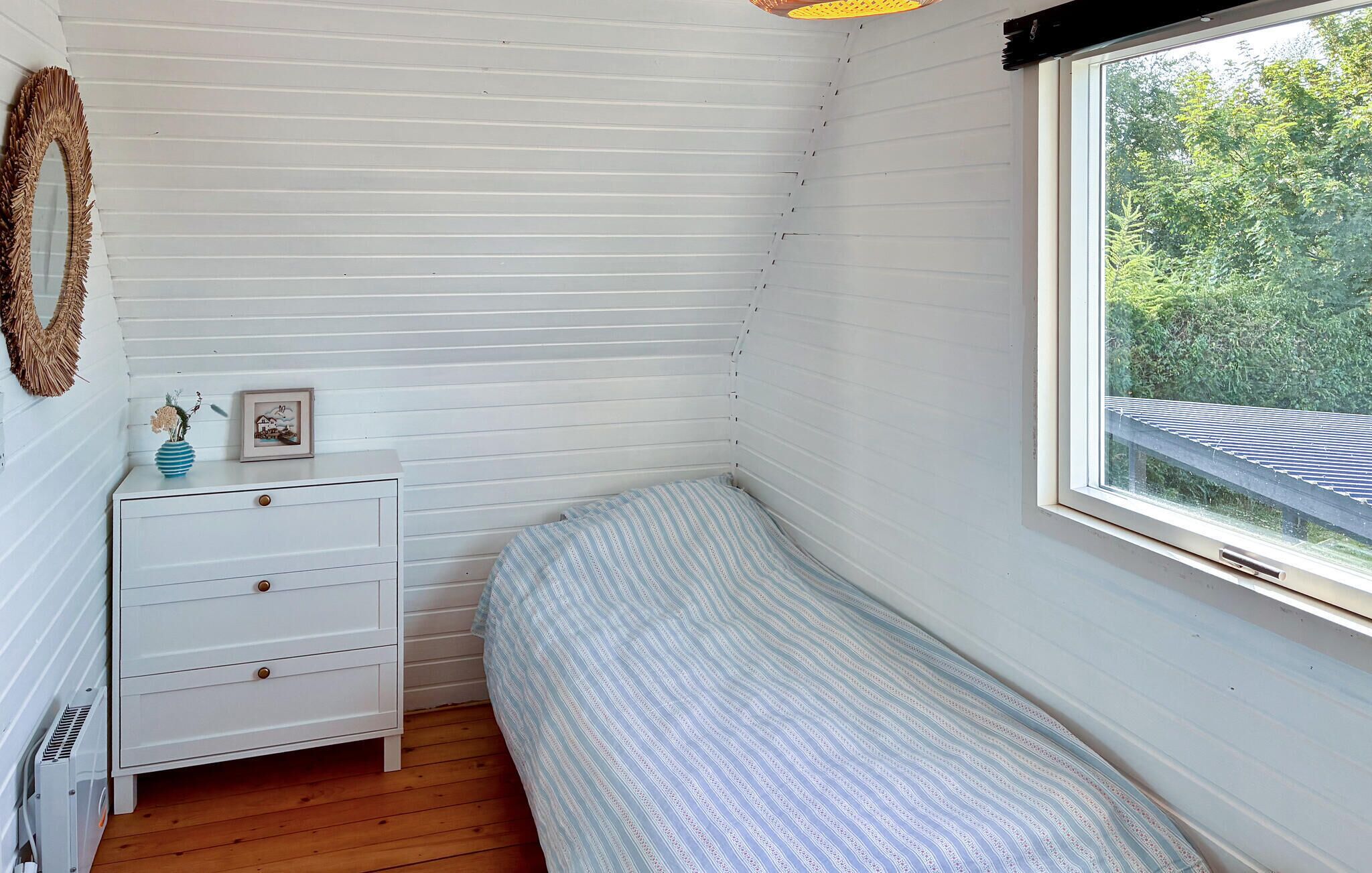 Quarto pequeno com uma cama de solteiro, janela, penteadeira branca com gavetas e um quadro em cima.
