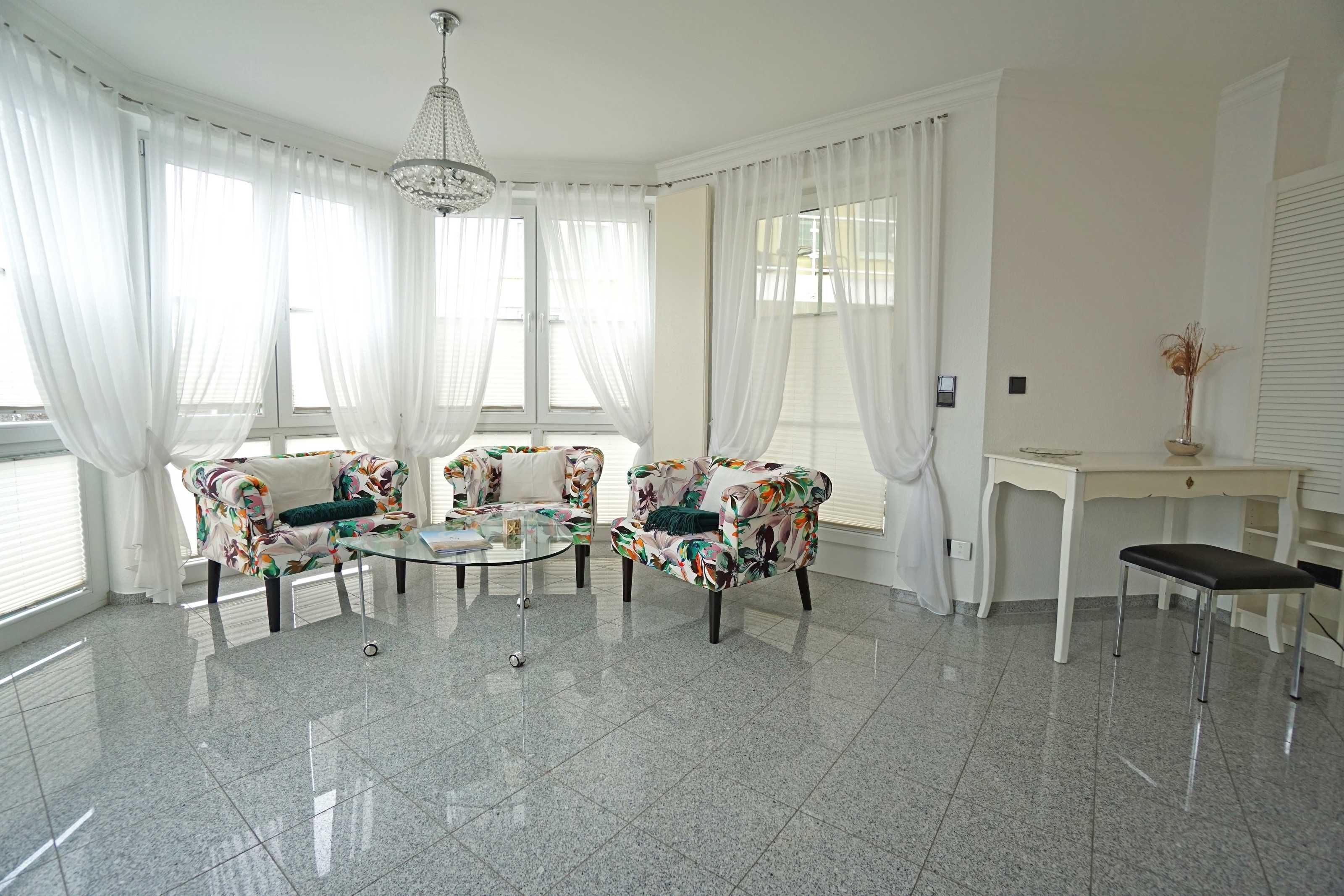 Una habitación con dos sillones de estampado floral, una mesa de centro de vidrio, un escritorio blanco y un banco.