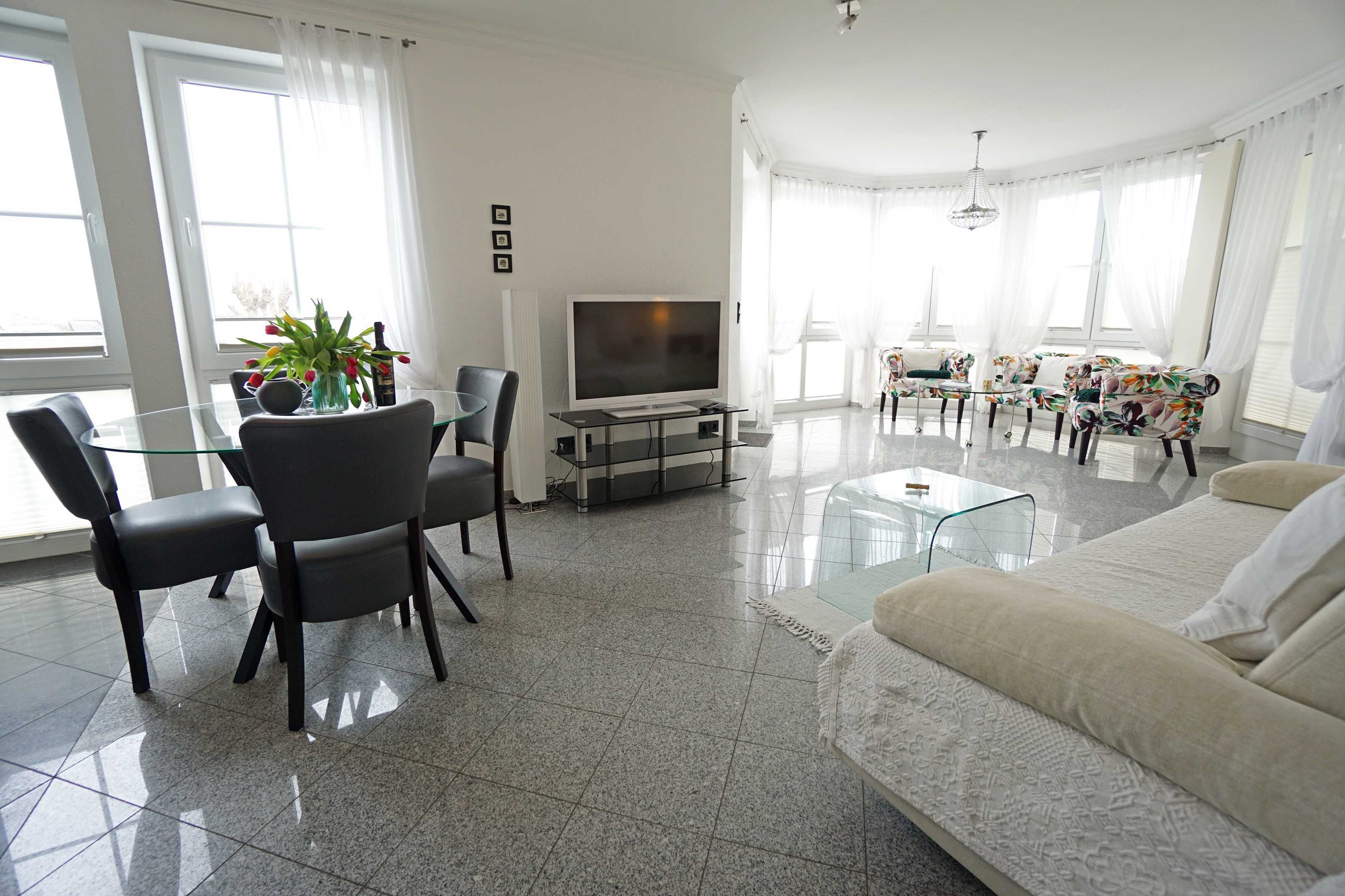 A modern living room with a glass dining table, black chairs, a beige sofa, and a flat-screen TV.