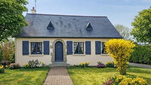 Een huis met twee verdiepingen, een donker dak, een centrale ingang met een deur en een pad van baksteen dat naar de voordeur leidt.