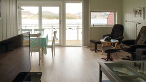 Amplia sala de estar con un gran ventanal corredero que da a un balcón con vistas a un cuerpo de agua y colinas. Cuenta con un comedor con mesa y sillas, una zona de estar acogedora con un sofá y sillones, y una pequeña mesa con un radio.