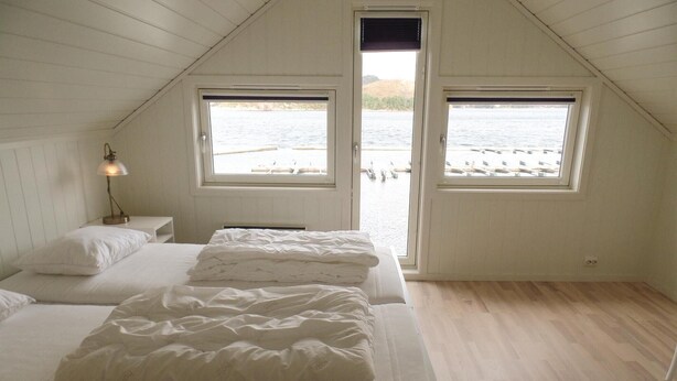Un dormitorio con dos camas, una mesita de noche con lámpara, y vista a un muelle y al agua a través de dos ventanas.