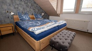 A bedroom with a sloped ceiling, a wooden bed frame, a grey tufted ottoman, a bedside table, a lamp, and a radiator. - (110) 3-room apartment - lake view - apartment complex Baltic Sea (Kühlungsborn)