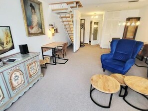 A living room with a blue armchair, a wooden coffee table, a dining table, and a staircase leading to another room. - (110) 3-room apartment - lake view - apartment complex Baltic Sea (Kühlungsborn)