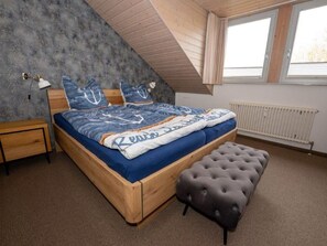 A bedroom with a sloped ceiling, a wooden bed frame, a grey tufted ottoman, a bedside table, a lamp, and a radiator.