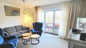A living room with a sofa, armchairs, a coffee table, a TV, and a balcony with curtains. - (110) 3-room apartment - lake view - apartment complex Baltic Sea (Kühlungsborn)