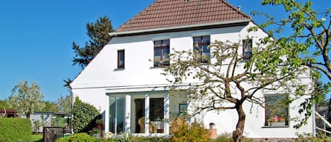 A white house with a brown roof, a garden with various plants, and a clear blue sky.
